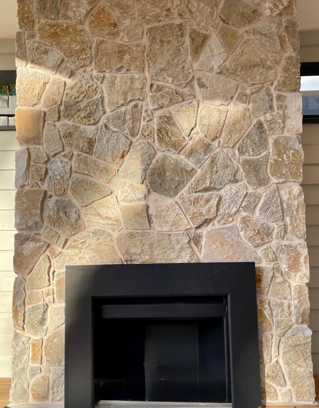 Natural stone feature wall with an irregular flagstone pattern in beige and warm grey tones, surrounding a modern black metal fireplace insert in a contemporary indoor living space.
