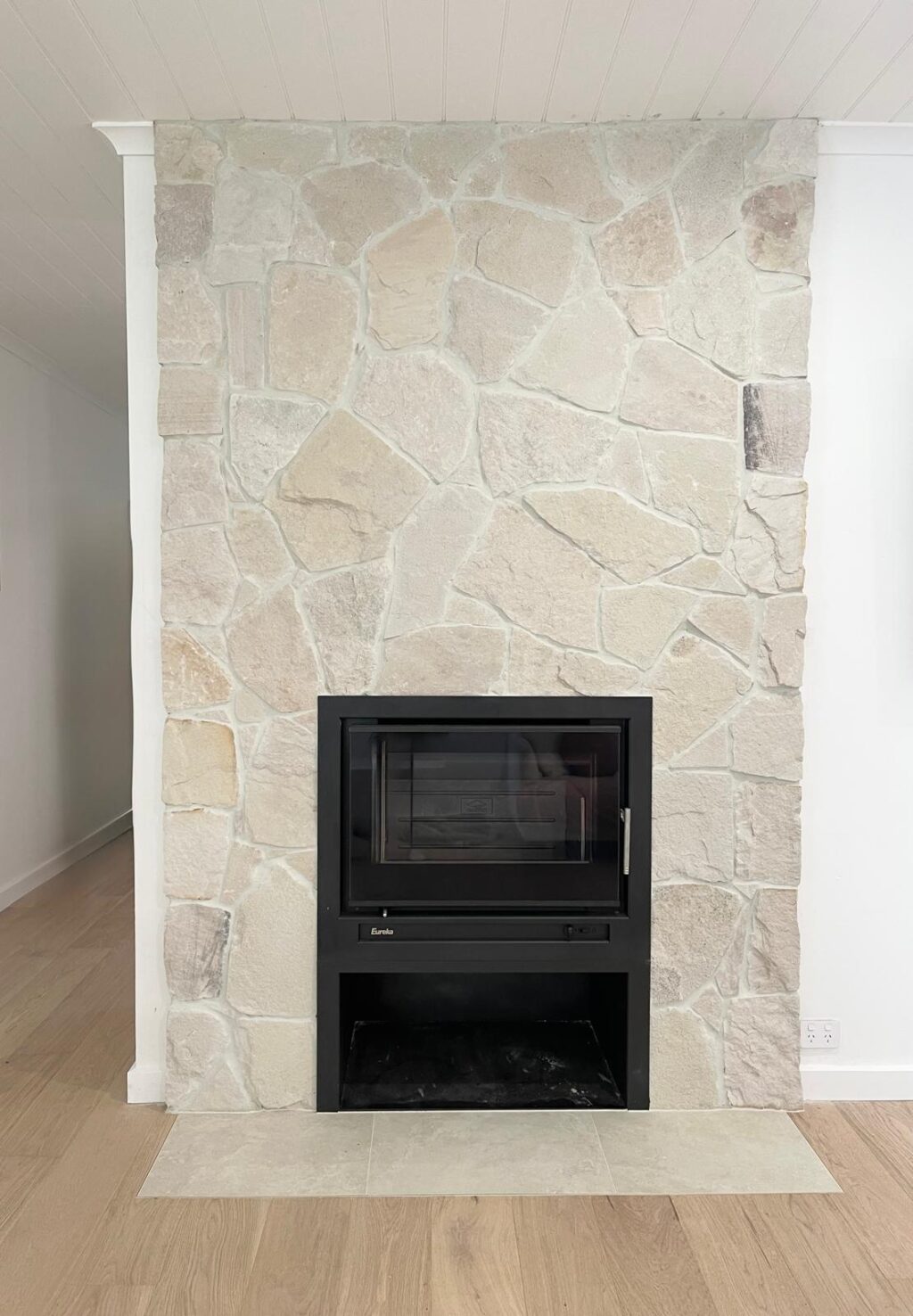 Interior feature wall made of Roman Sandstone with a built-in black wood-burning fireplace, complemented by light timber flooring and a white panelled ceiling.