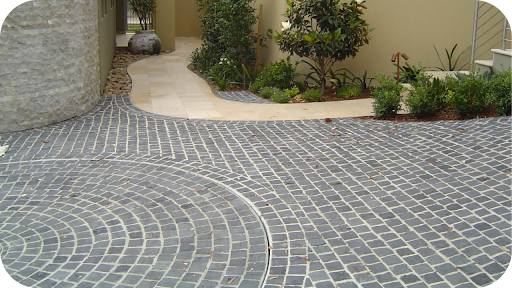 A curved path and circular driveway made of dark gray cobblestone pavers meanders through a lush garden.