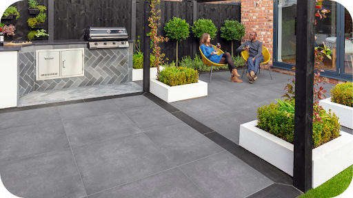 A couple relaxing in yellow chairs on a stylish gray-paved patio with a black privacy fence, an outdoor kitchen, and potted plants.