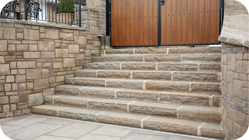 A wide flight of outdoor steps made from rectangular stone blocks leads up to a modern wooden gate, with a matching stone retaining wall on the left.