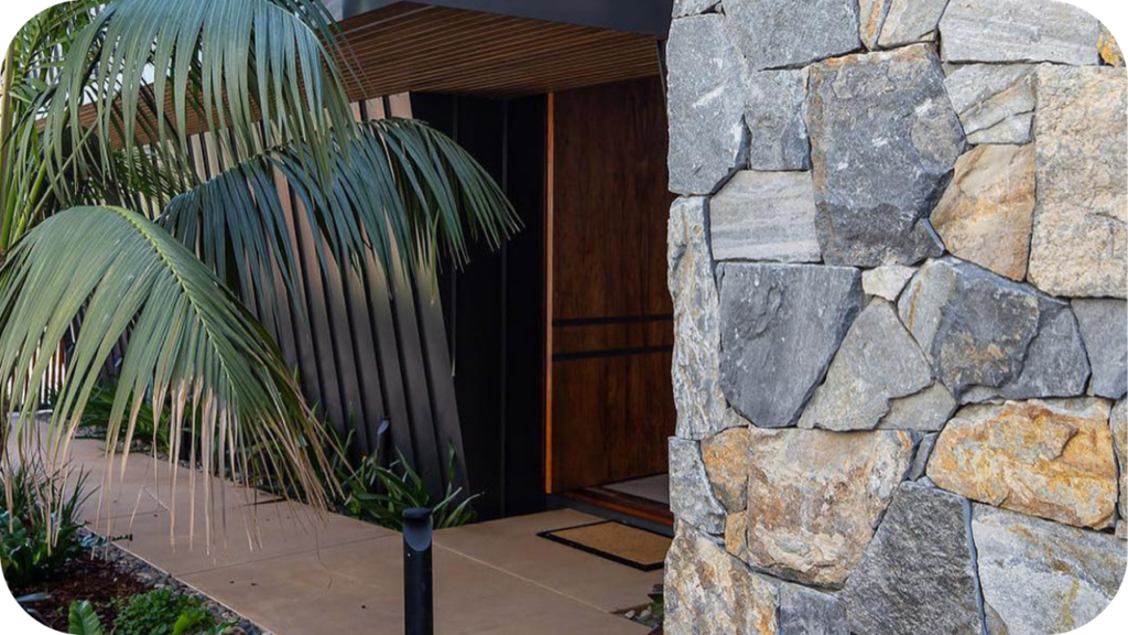 Close-up of rugged stone cladding on a modern entryway, framed by lush palm leaves and sleek vertical paneling.
