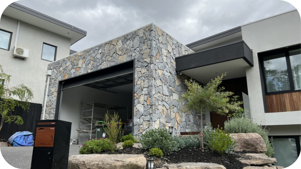 Textured stone cladding highlights a modern garage facade, blending with native garden landscaping and boulder edging.