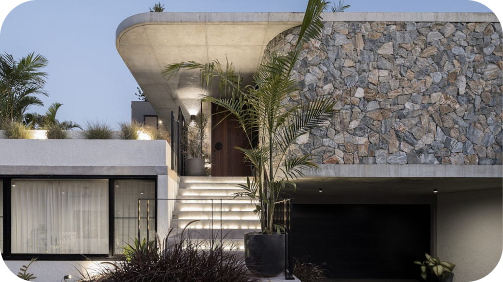 Contemporary house facade with raw stone cladding, curved concrete roofline, palm landscaping and front-lit stairs.