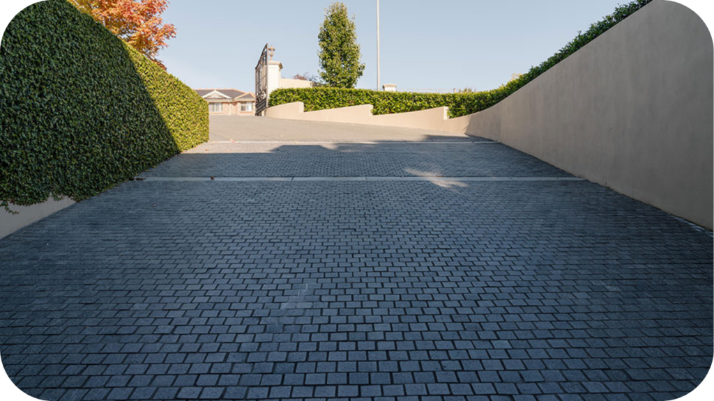 Driveway paved with dark cobblestones, framed by tall manicured hedges and soft curved walls in a quiet Australian suburb.