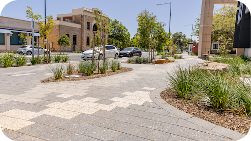 Stone-paved Australian footpath with native plantings, curved garden beds and traffic passing heritage sandstone buildings.