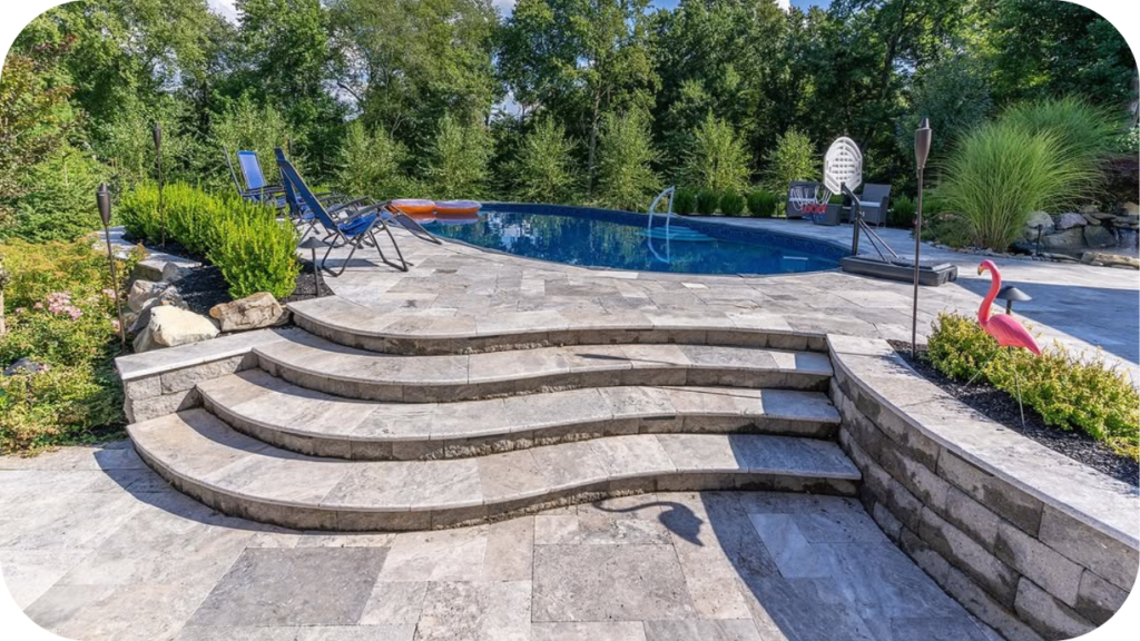 Curved stone steps and poolside area paved with travertine tiles, surrounded by lush greenery and lounge chairs for outdoor relaxation.