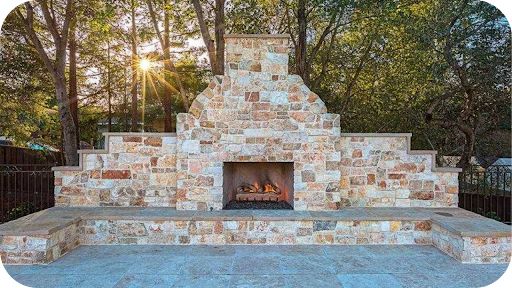 Outdoor stone fireplace with warm cladding and stepped design, glowing with firelight among tall trees in a garden setting
