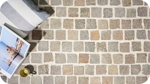 Square porphyry cobblestones in warm grey and brown shades, styled with a cushion, book, and coffee cup, creating a cosy and inviting outdoor setting.