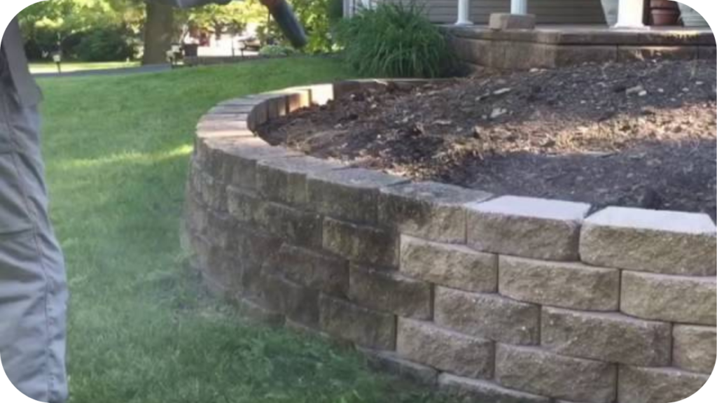 Person cleaning a curved stone retaining wall beside a lawn, highlighting easy maintenance and long-lasting durability of stone.