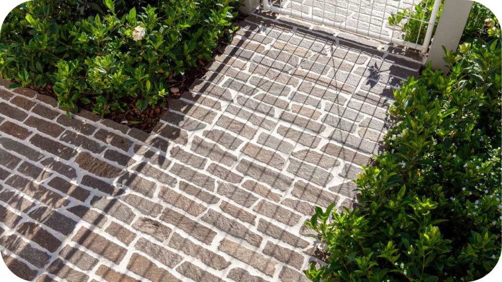 Walkway paved with filetti porphyry pavers laid in a fan pattern, framed by flowering hedges and a sunlit white entry gate.