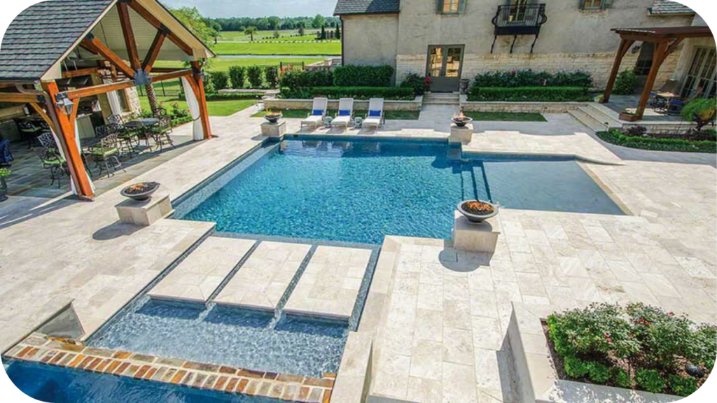 Luxury backyard pool surrounded by light travertine tiles, sun loungers, and shaded patio seating, blending elegance with outdoor comfort.
