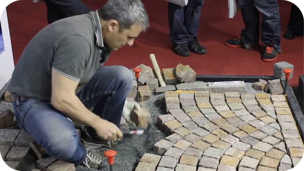  Man installing porphyry cobblestones in a curved pattern, carefully setting each stone on a compacted base during a live demonstration of traditional paving techniques.