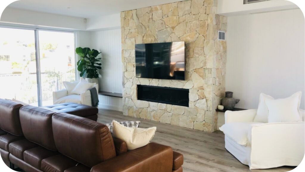 Bright living room with cream quartz wall cladding around a TV and fireplace, paired with leather and white armchairs.