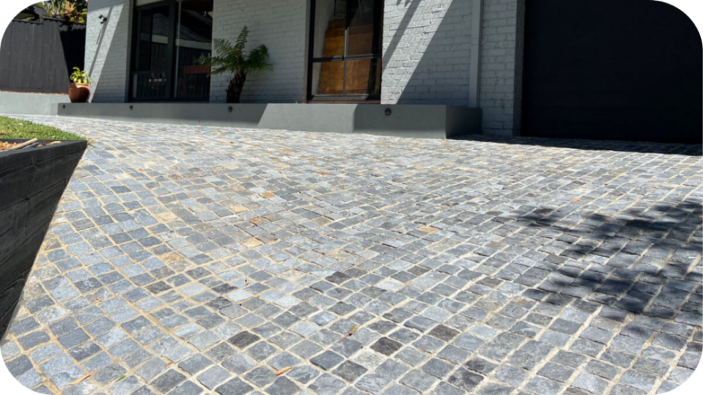 A wide, grey cobblestone driveway in front of a modern house with white-painted brick walls and a raised porch.
