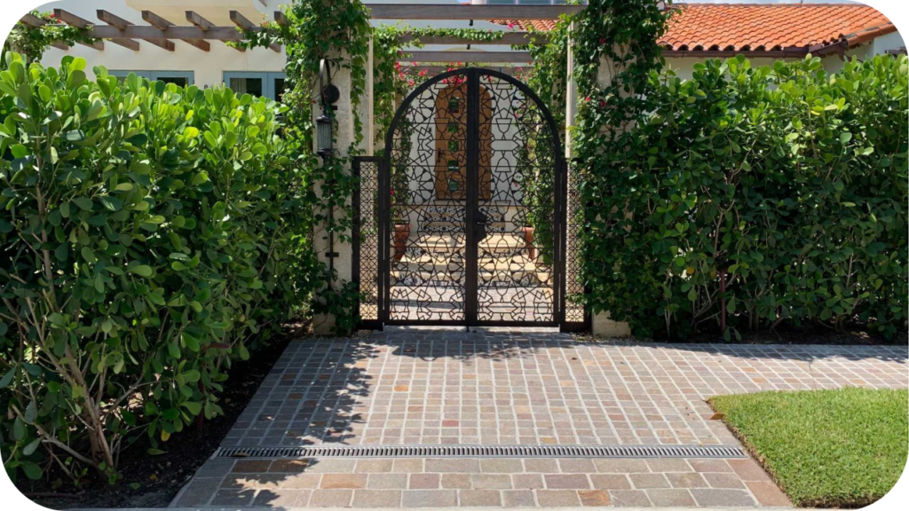 A paver walkway leads through green hedges to an ornate, arched metal gate set between two vine-covered pillars.
