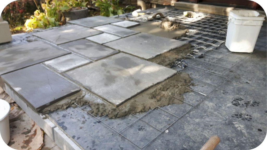 Square stone pavers being laid on a thick bed of wet mortar over a wire mesh grid during a patio installation.
