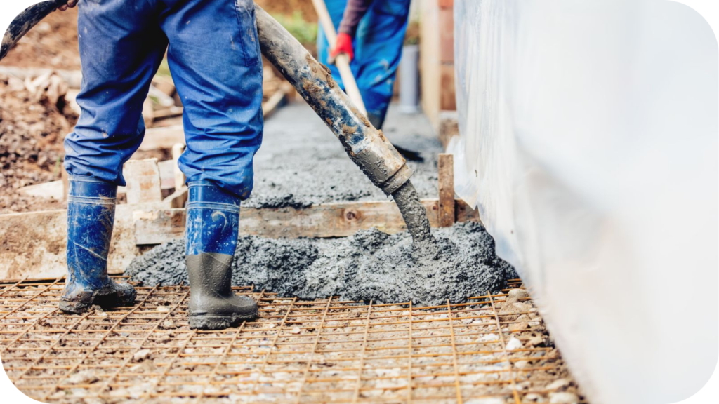A worker in blue rubber boots stands on a rebar grid as wet concrete is poured from a large pump hose into a foundation.