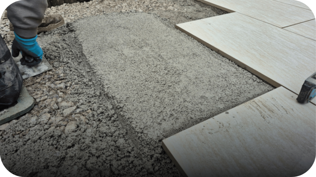 A worker kneels to level a bed of sand or mortar with a trowel while laying large paver stones for a patio.