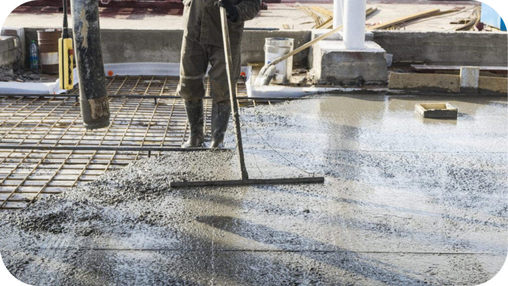 A worker in rubber boots uses a long-handled bull float to level a large, wet concrete slab being poured over a rebar grid.