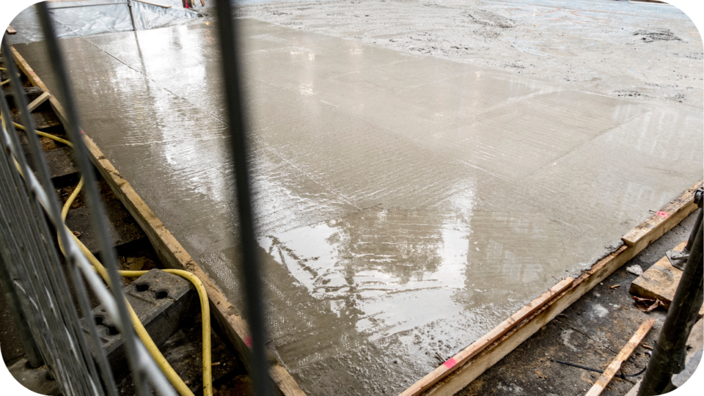 A freshly poured, wet concrete slab with a reflective surface, enclosed by wooden formwork on a construction site.