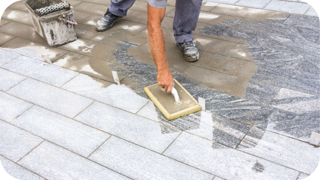 A worker uses a sponge float to apply wet grout into the joints of newly laid stone paver tiles.