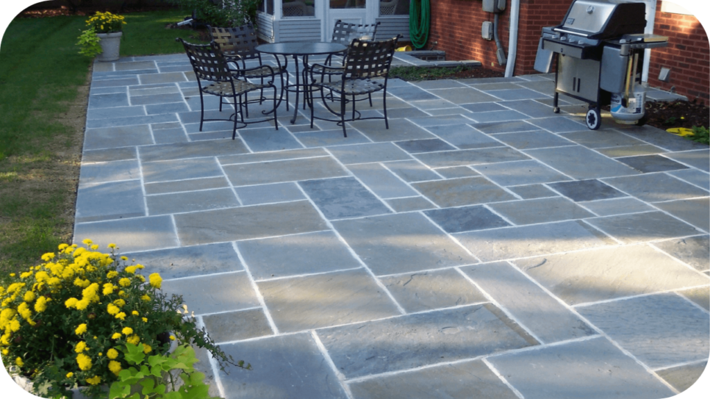 Backyard dining area with bluestone pavers, metal table and chairs, and vibrant yellow flowers adding charm and style.
