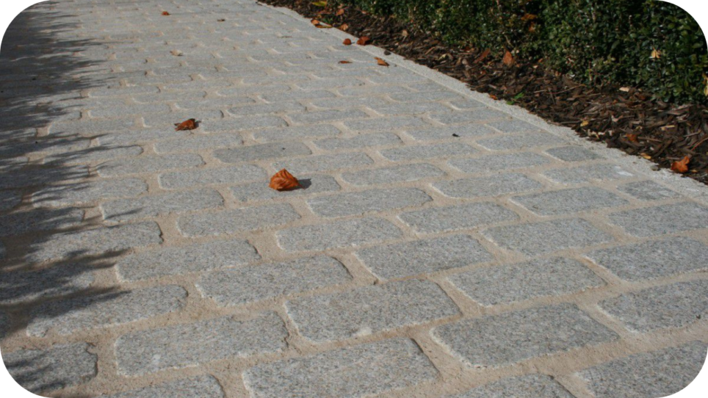 Outdoor pathway paved with rectangular grey granite cobblestones, framed by garden mulch and autumn leaves.