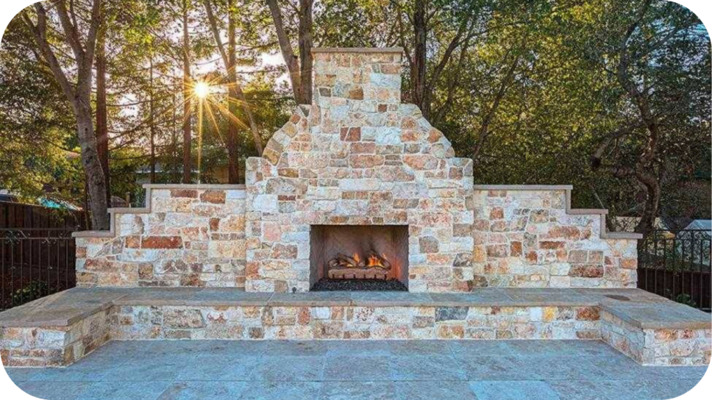 Outdoor natural stone fireplace with warm-toned cladding and stepped design, blending seamlessly with surrounding trees and evening sunlight.
