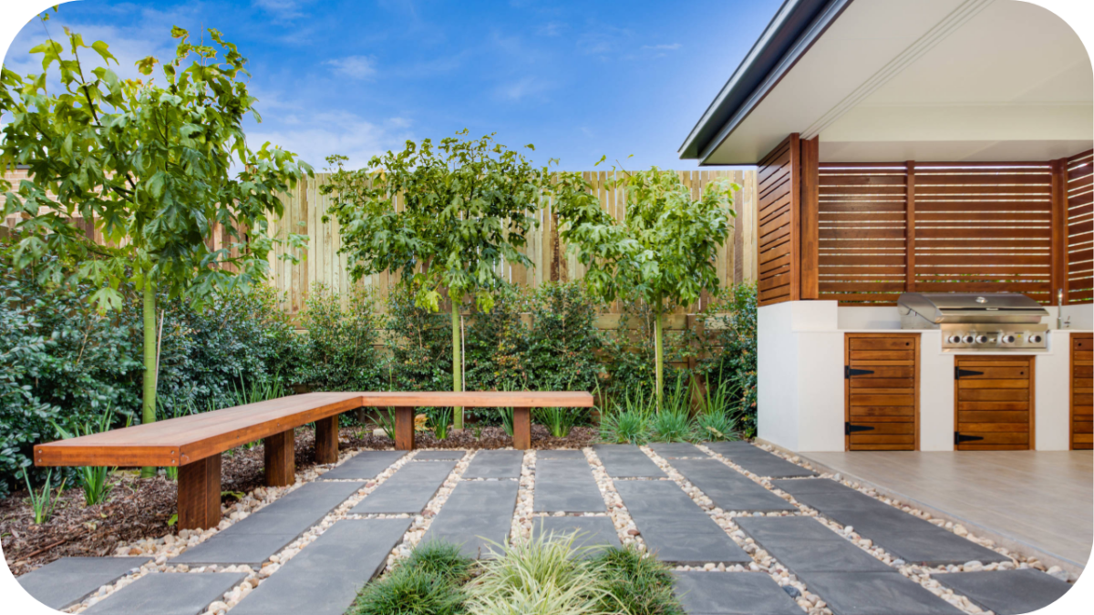 Outdoor entertaining area with stone paving, timber benches, and custom joinery BBQ setup surrounded by greenery, showing functional landscape design.