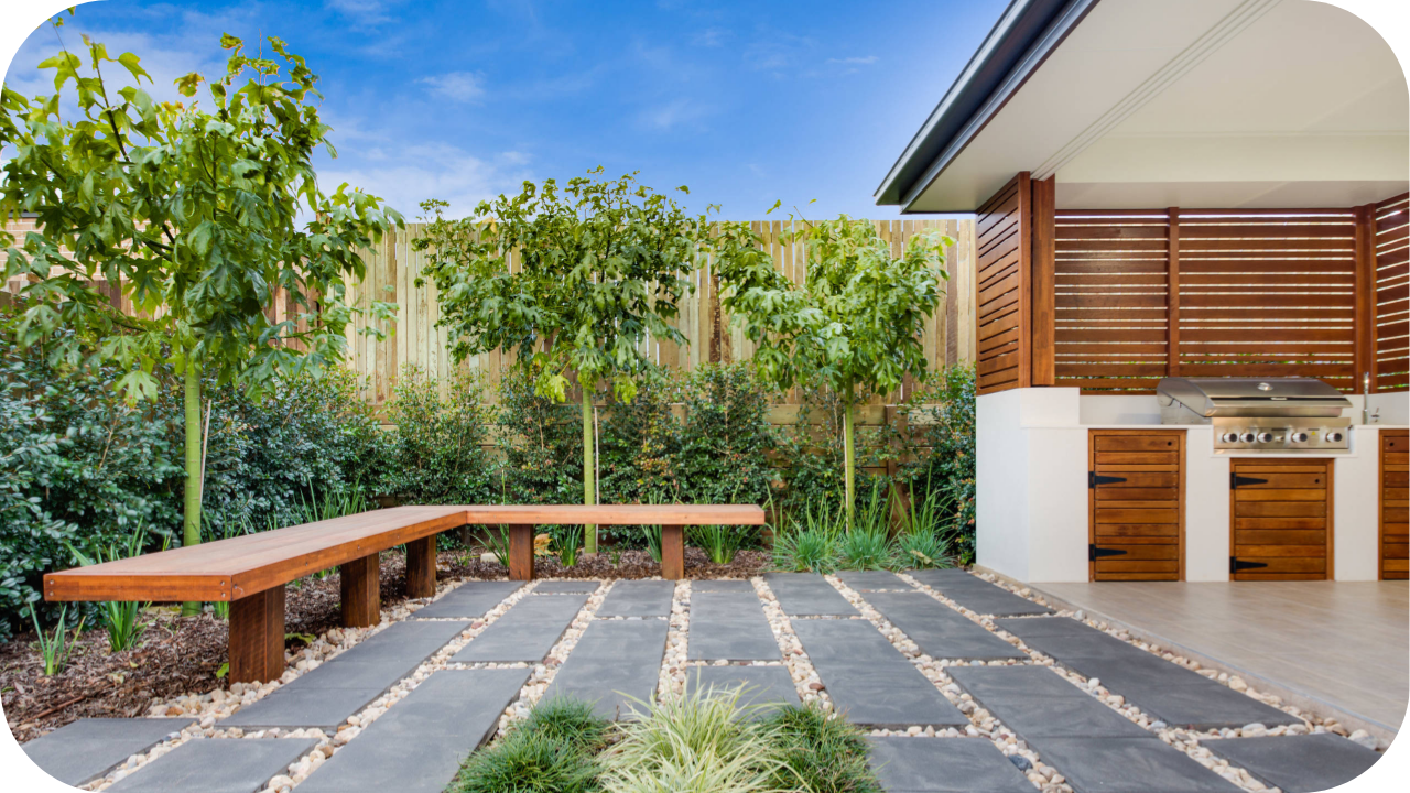 Outdoor entertaining area with stone paving, timber benches, and custom joinery BBQ setup surrounded by greenery, showing functional landscape design.