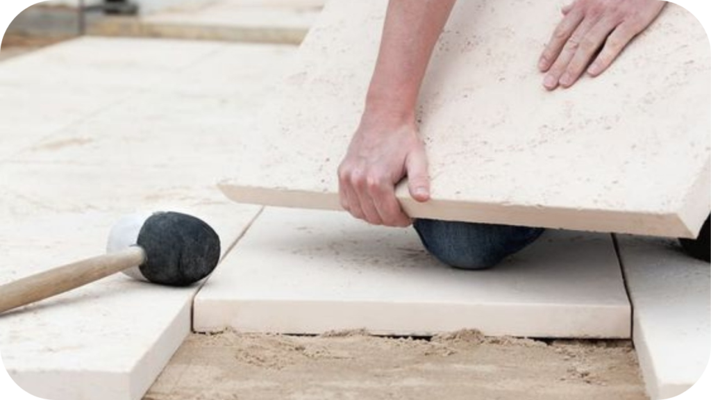 Hands placing a light coloured stone paver onto a sand base with a mallet nearby, showing a simple DIY paving step in progress. 