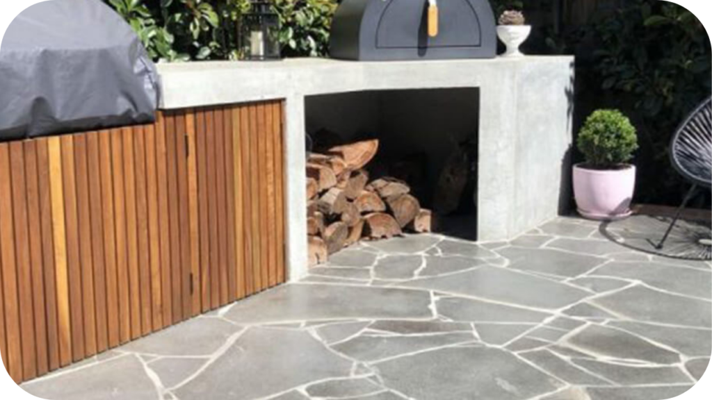 Random crazy paving in natural grey stone surrounding an outdoor kitchen with timber cabinetry and a pizza oven, creating a relaxed modern entertaining space.