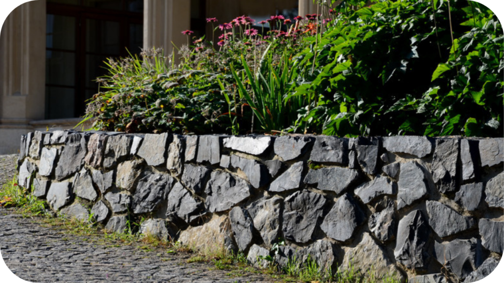 Natural stone retaining wall built with irregularly shaped stones supporting a raised garden bed filled with greenery and flowers near a home.