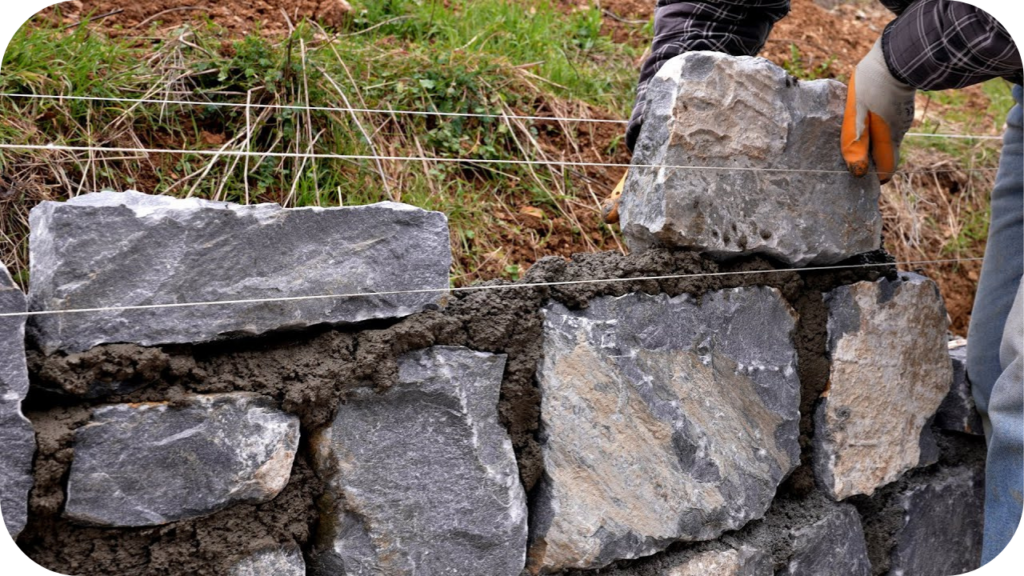Two people laying large natural stone blocks with mortar and string lines to build a stable retaining wall on a sloped outdoor site.